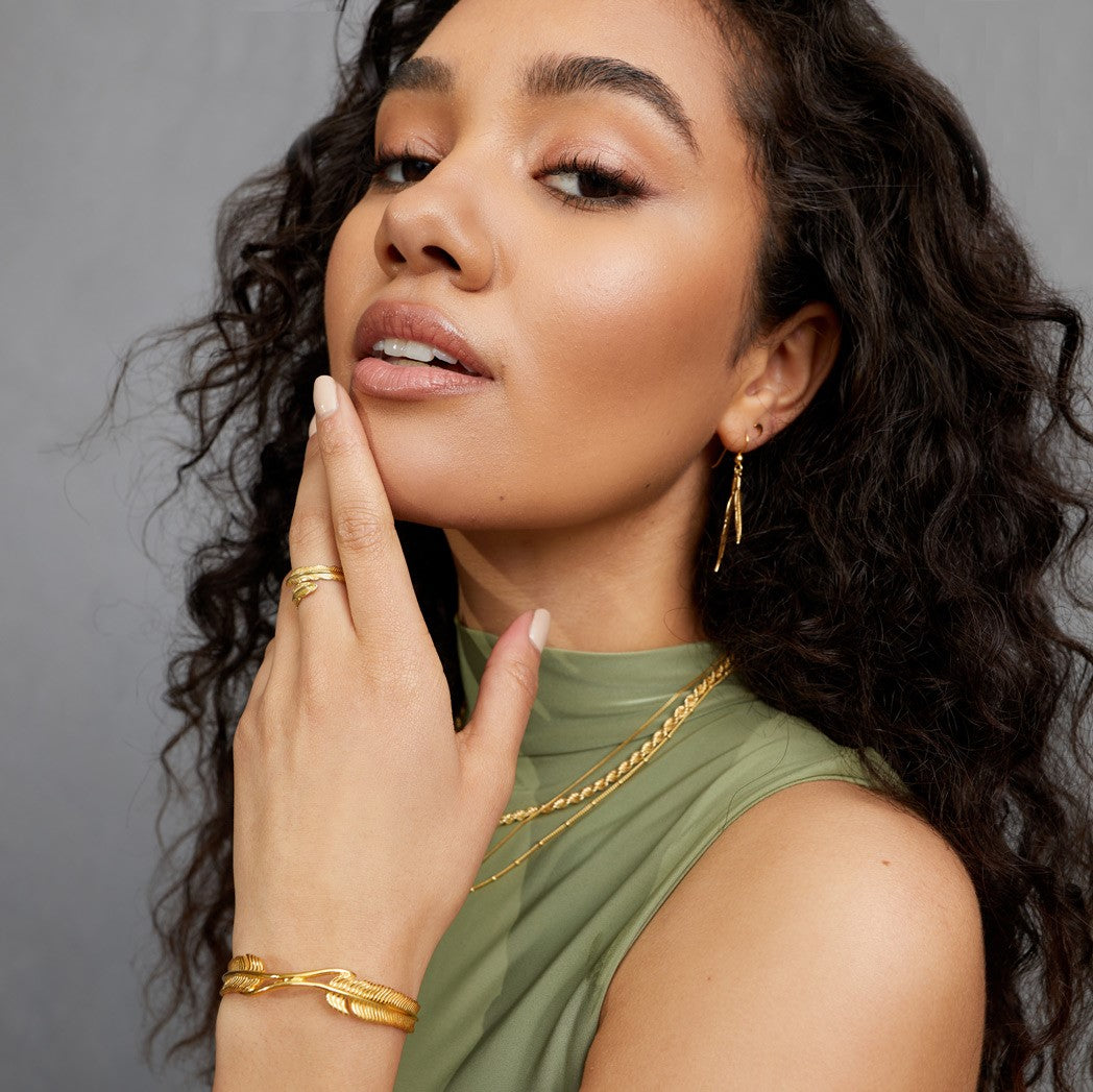 A close up of a woman wearing a green top whilst holding her hand up to her face, wearing a FIYAH Yellow Gold Plume Bangle, a FIYAH Yellow Gold Adjustable Plume Ring, and a pair of FIYAH Yellow Gold Dual Plume Earrings