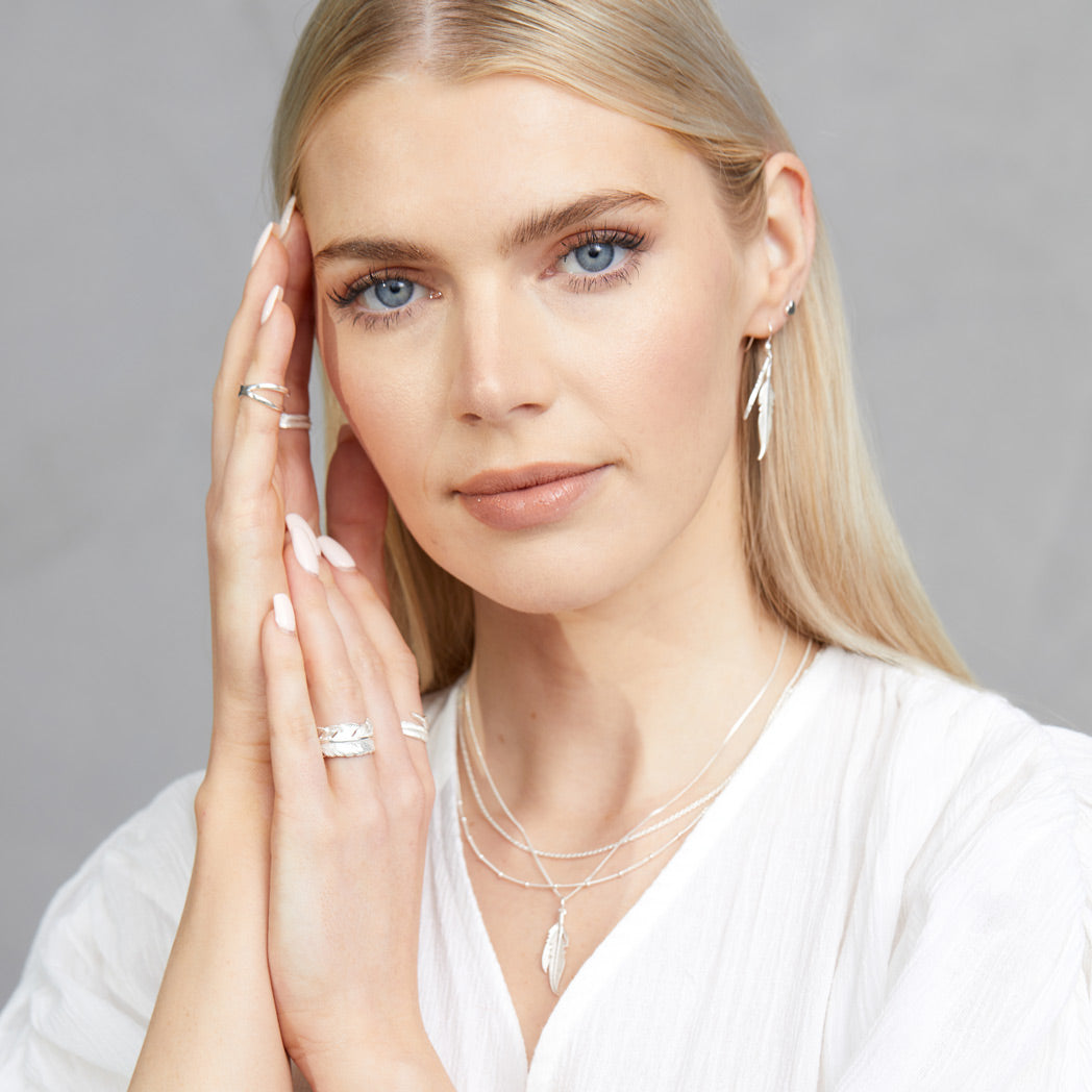 A headshot of a woman holding her hand up close to her face, whilst wearing a FIYAH Silver Adjustable Plume Ring, a FIYAH Silver Imperial Plume Ring, a pair of FIYAH Silver Dual Plume Earrings and a FIYAH Silver Dual Plume Necklace