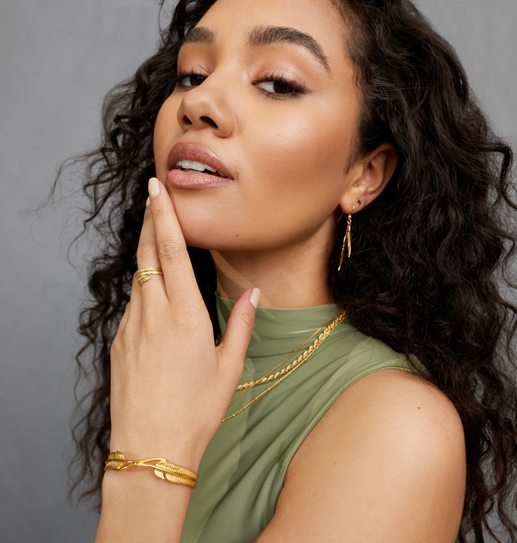 A close up of a woman in a green top, holding her hand up to her face, wearing a FIYAH Yellow Gold Plume Bangle, a FIYAH Yellow Gold Adjustable Plume Ring, and FIYAH Yellow Gold Dual Plume Earrings