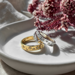Two rings, one gold plated and one sterling silver, on a white plate with flowers in the background.