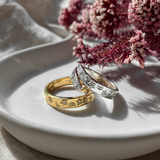 Two rings, one gold plated and one sterling silver, on a white plate with flowers in the background.