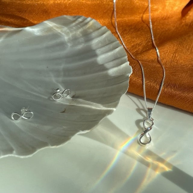 FIYAH Silver Infinity Stud Earrings on a shell, next to the FIYAH Silver Infinity Necklace lay on a piece of orange material